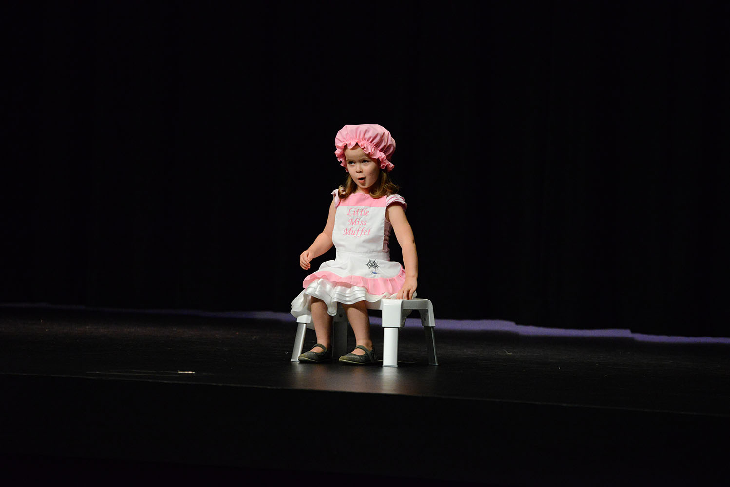 Very young speech & drama performer. Sitting on a stool on stage wearing a white and pink costume.