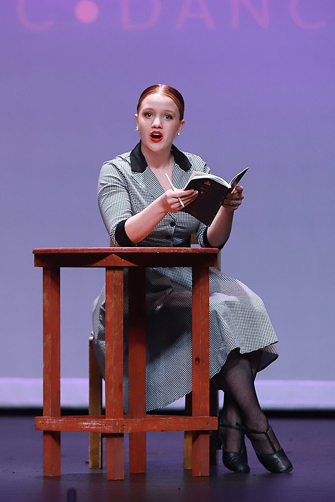 Teenaged Song & dance performer onstage, sitting on a stool leaning on a small table with a book open.